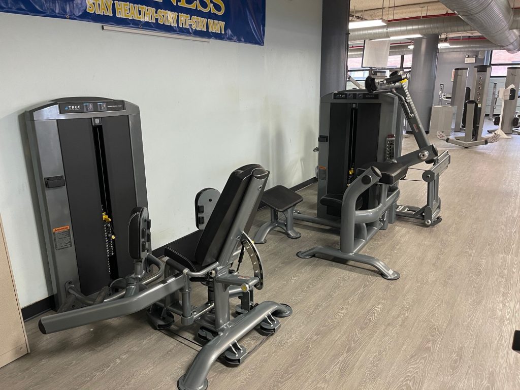 Two strength training machines in a gym with gray flooring and light-colored walls. A blue and yellow fitness banner hangs above. Other gym equipment is visible in the background.