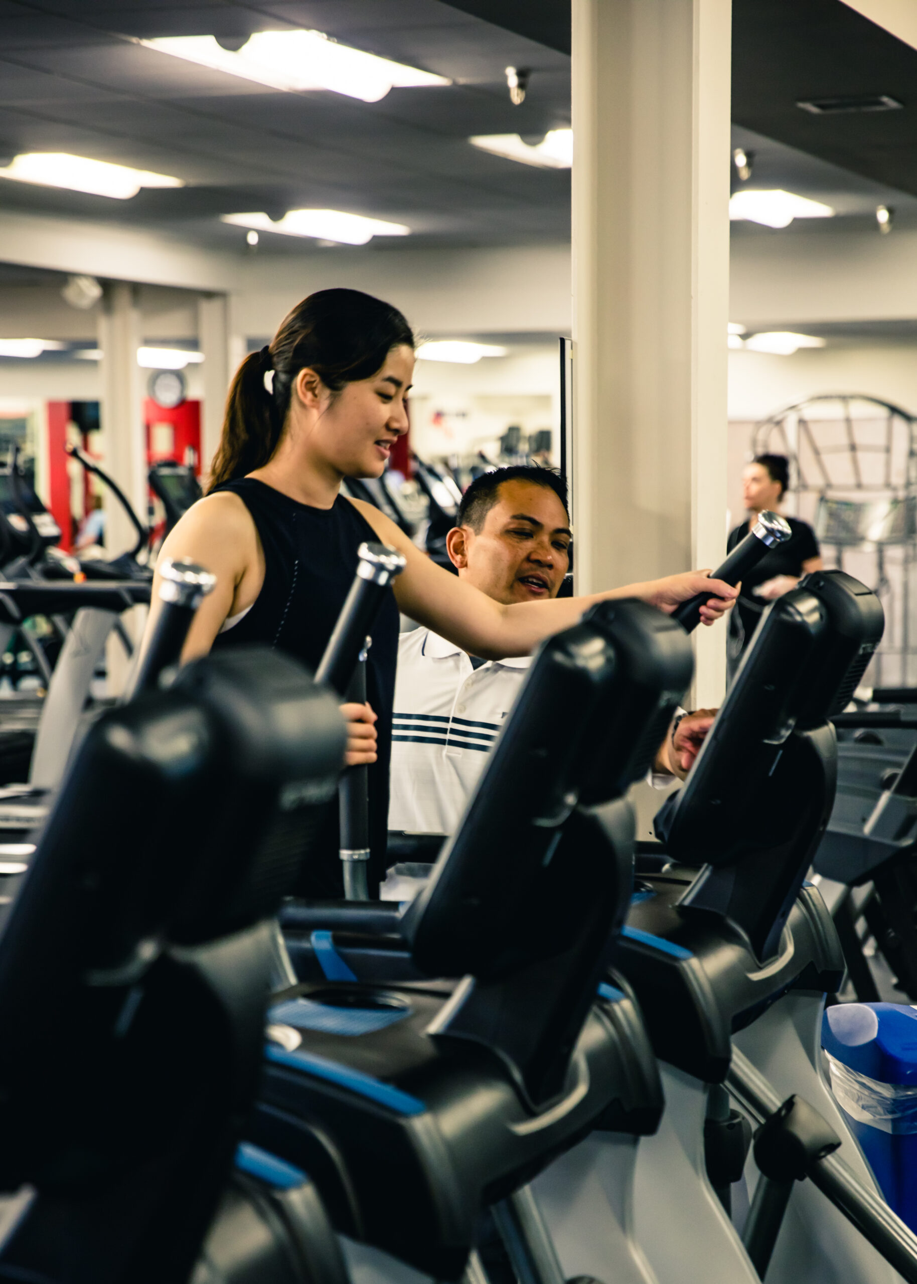 A woman and a man stand next to exercise machines in a gym. The woman is pointing at the screen of an elliptical trainer while the man watches, surrounded by other gym equipment.