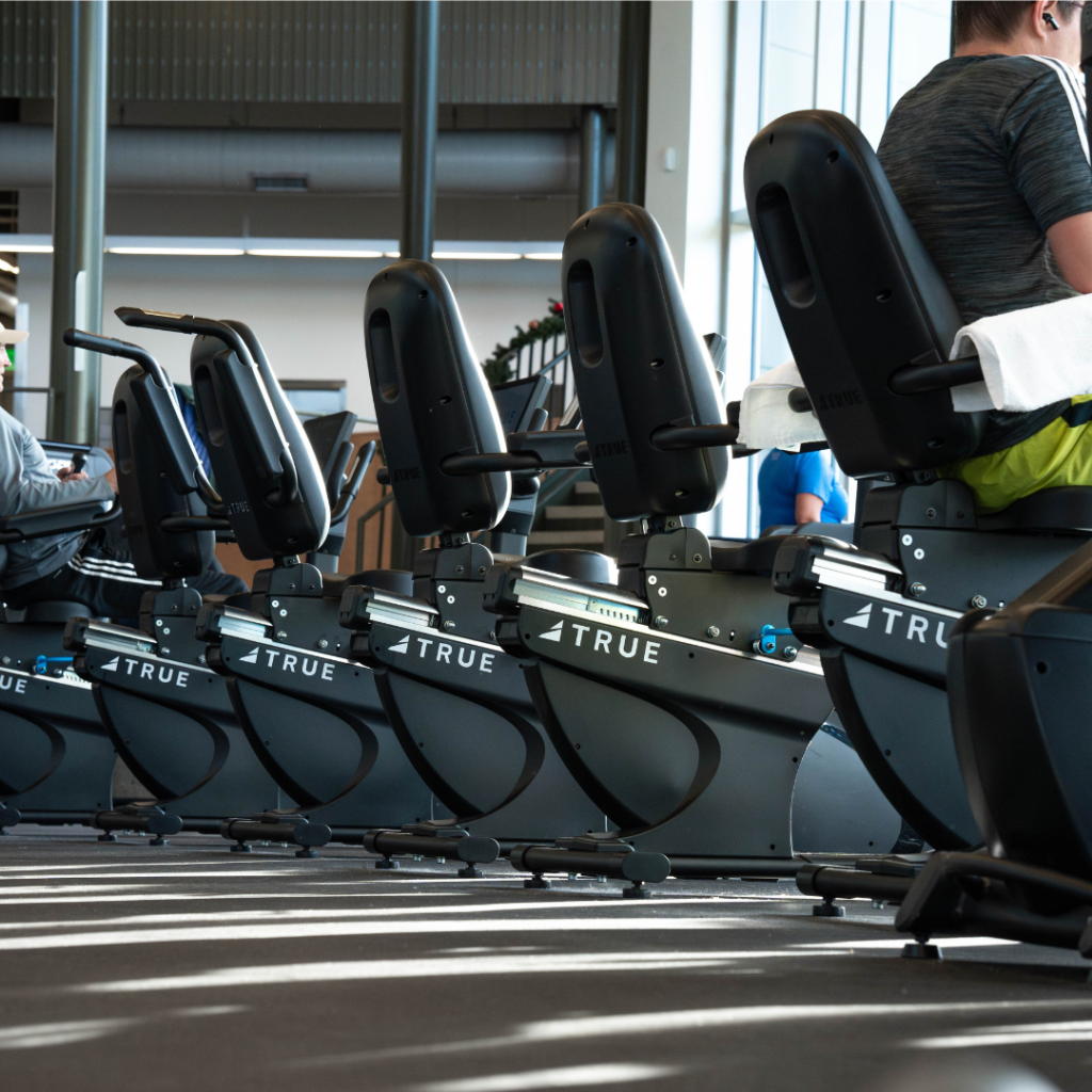 A row of stationary exercise bikes in a gym, with people sitting on them and towels draped over the backs of some seats. The gym has large windows, letting in natural light.