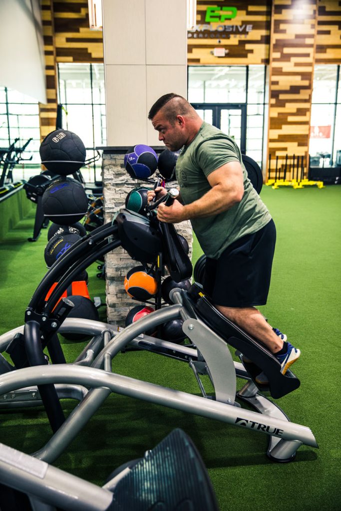 A man in a green t-shirt exercises on a step machine in a gym, surrounded by colorful medicine balls and fitness equipment on green turf flooring.
