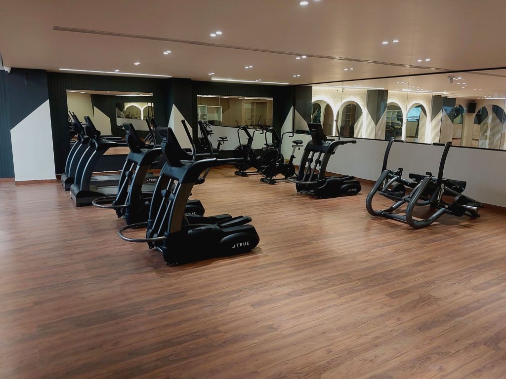 A modern gym with several black elliptical machines and exercise bikes on a polished wooden floor, large mirrors on the wall, and bright ceiling lights creating a spacious and clean atmosphere.