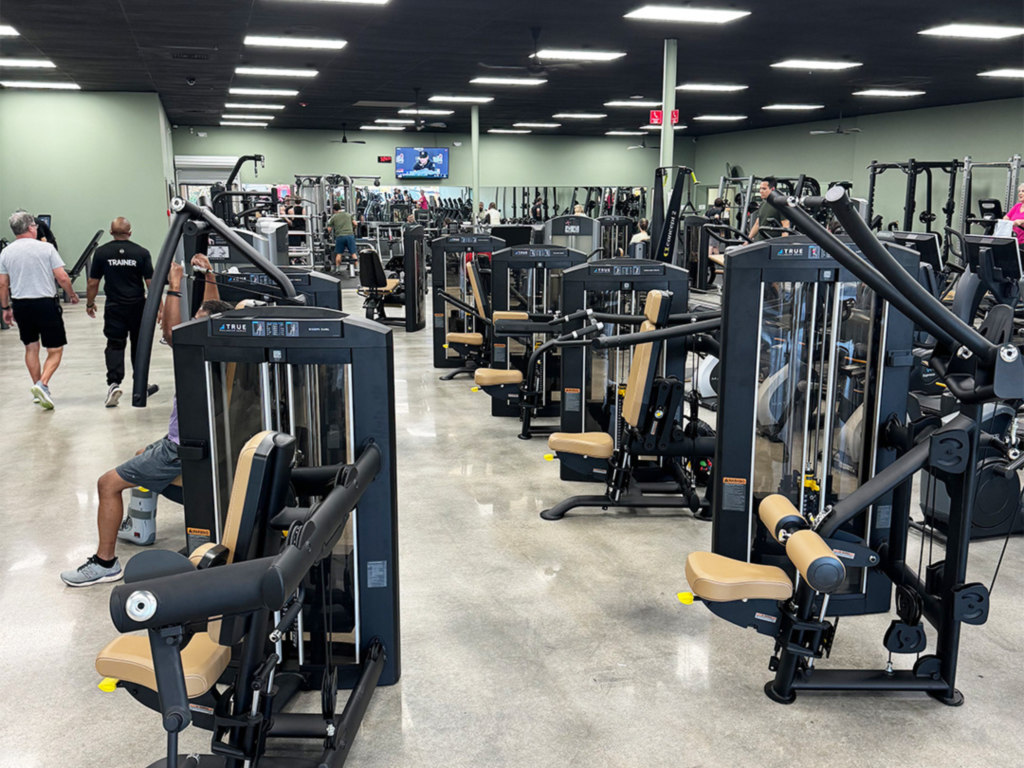 A spacious gym with multiple strength training machines, some in use, and people walking or exercising. The floor is polished, walls are light green, and ceiling lights are bright. A TV is mounted on the far wall.