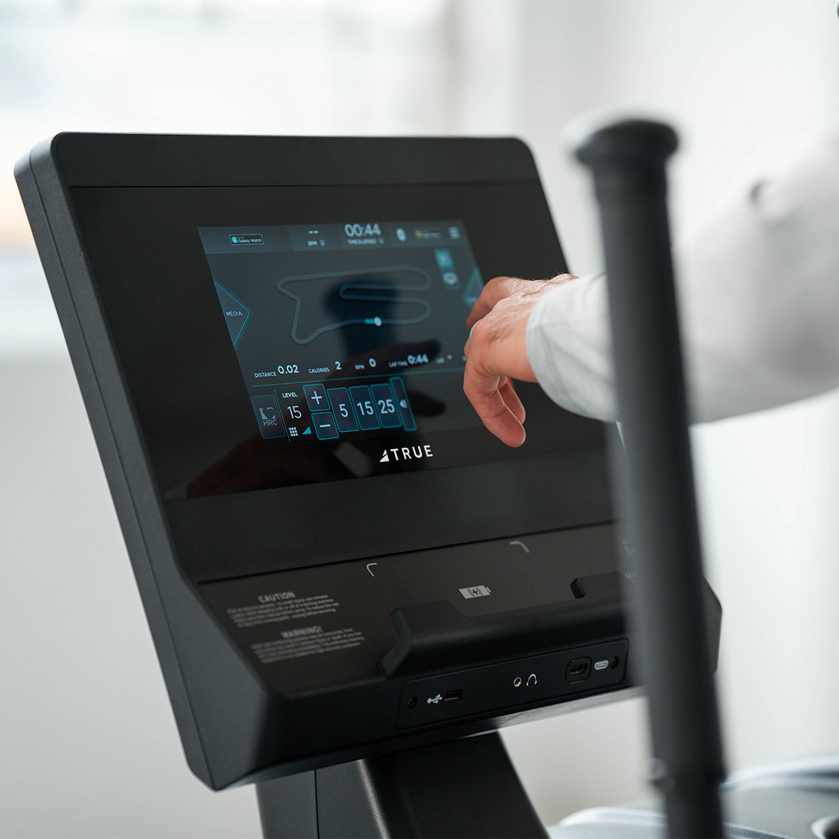 A person’s hand interacts with the touchscreen display of a fitness machine, adjusting workout settings. The screen shows exercise options and data, with a handlebar visible in the foreground.