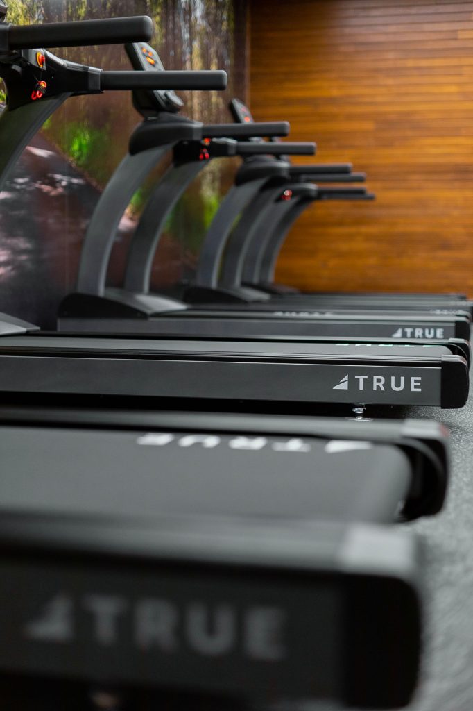 A row of black TRUE treadmills lined up in a gym with a wood-paneled wall and a nature-themed mural in the background.