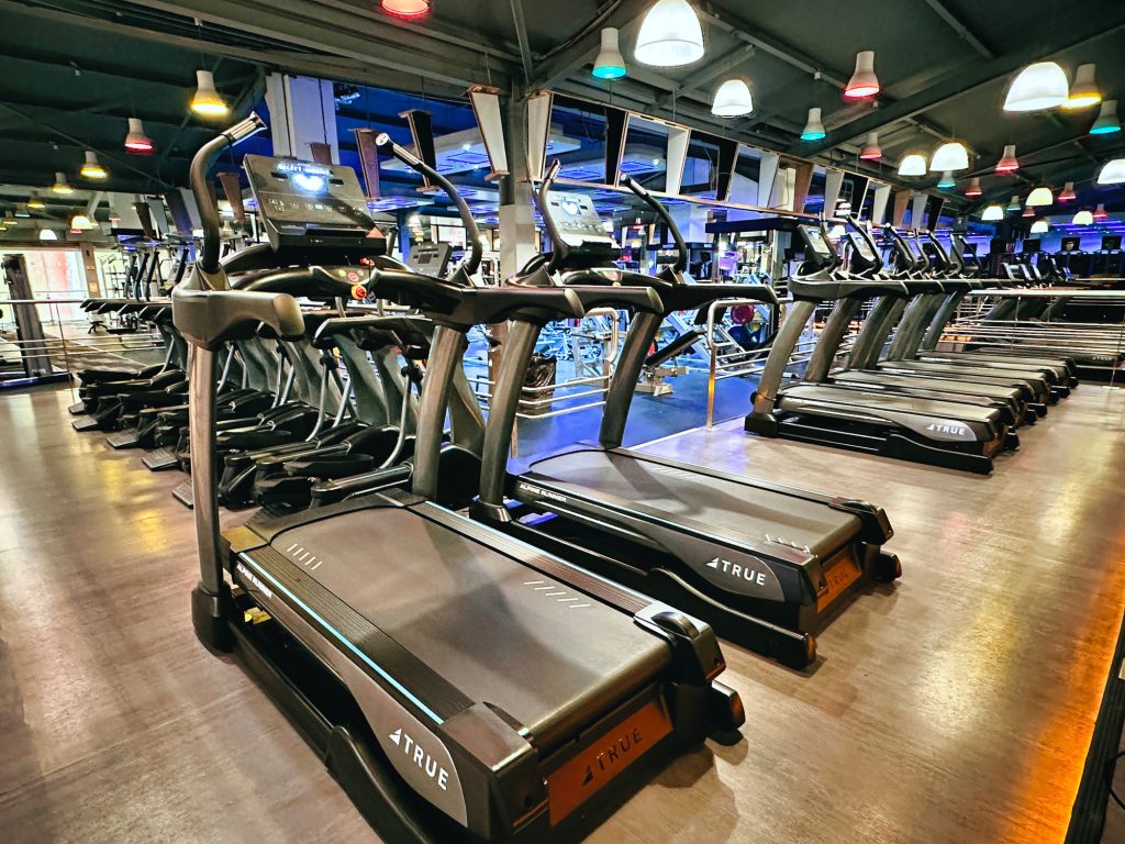 A row of treadmills in a modern gym with mirrored walls and colorful ceiling lights, reflecting fitness equipment and creating a bright, energetic atmosphere.
