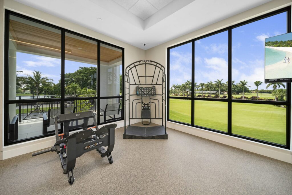 A sunlit indoor sports room with large windows overlooking a green lawn and palm trees, featuring a baseball pitching machine and a baseball rebound net. A TV in the corner displays a tropical beach scene.