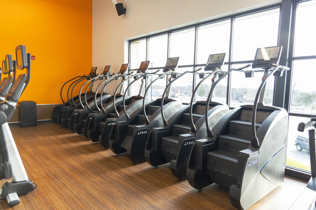 A row of stair climbing machines lines a gym with wood flooring, orange and white walls, and large windows letting in natural light. Each machine has a digital display screen.