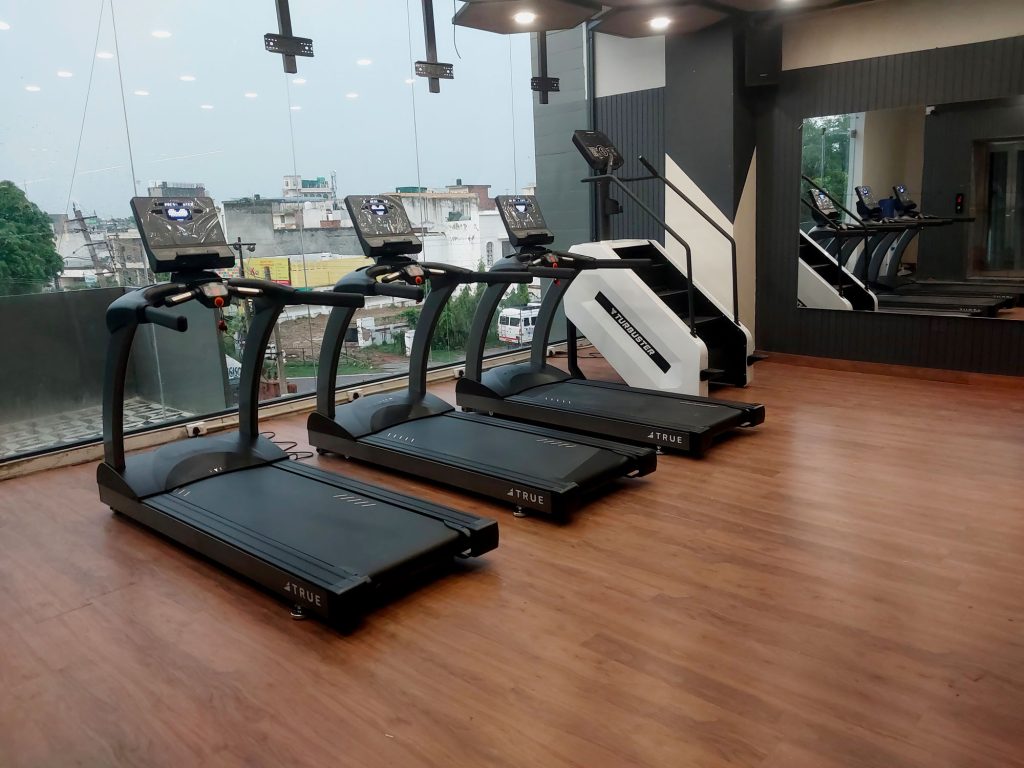 A row of four modern treadmills facing large windows and mirrors in a spacious gym with wooden flooring, overlooking buildings and greenery outside.
