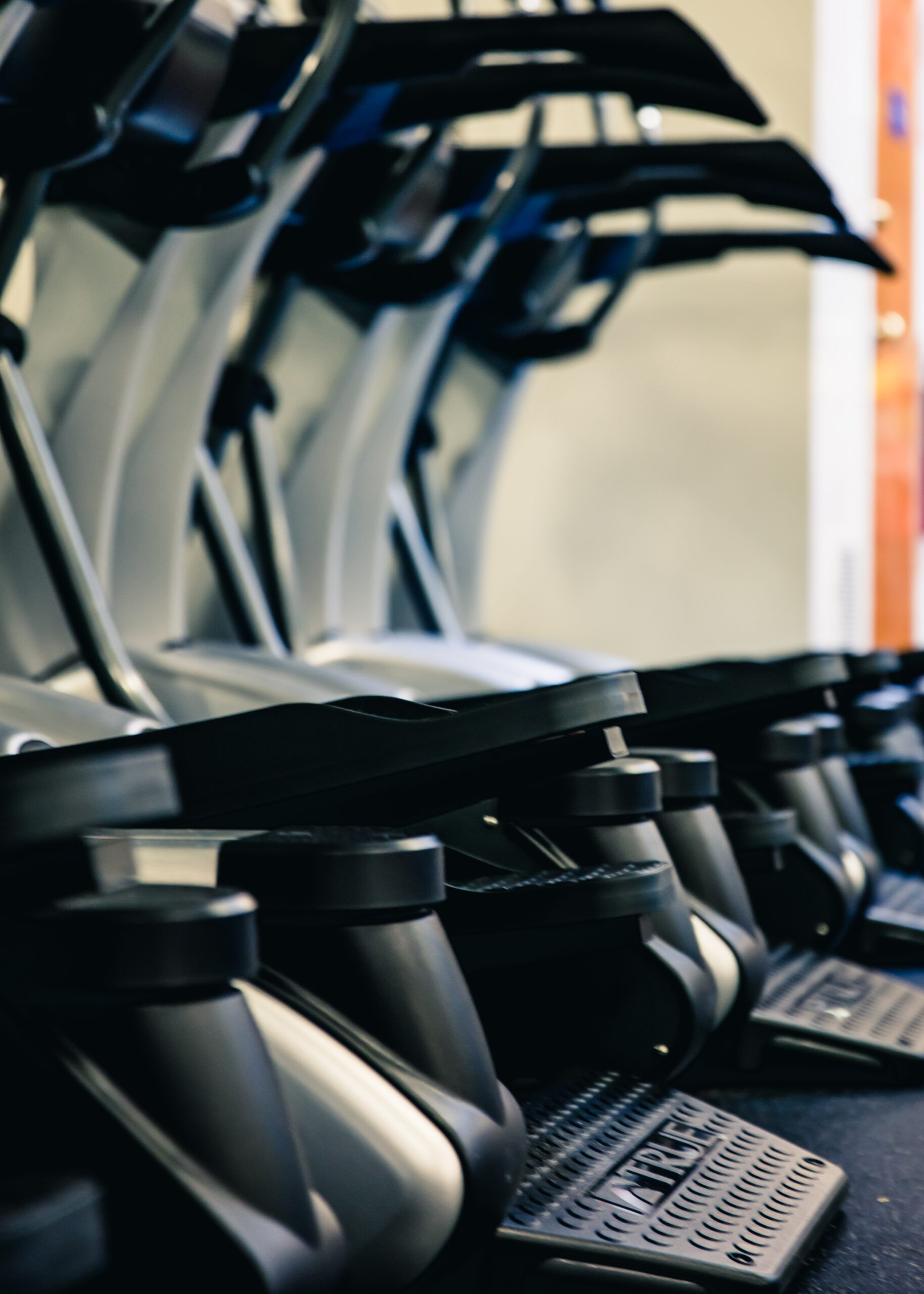 A row of treadmills in a gym, angled closely together, with handlebars and control panels visible. The background is softly blurred, emphasizing the exercise equipment in the foreground.