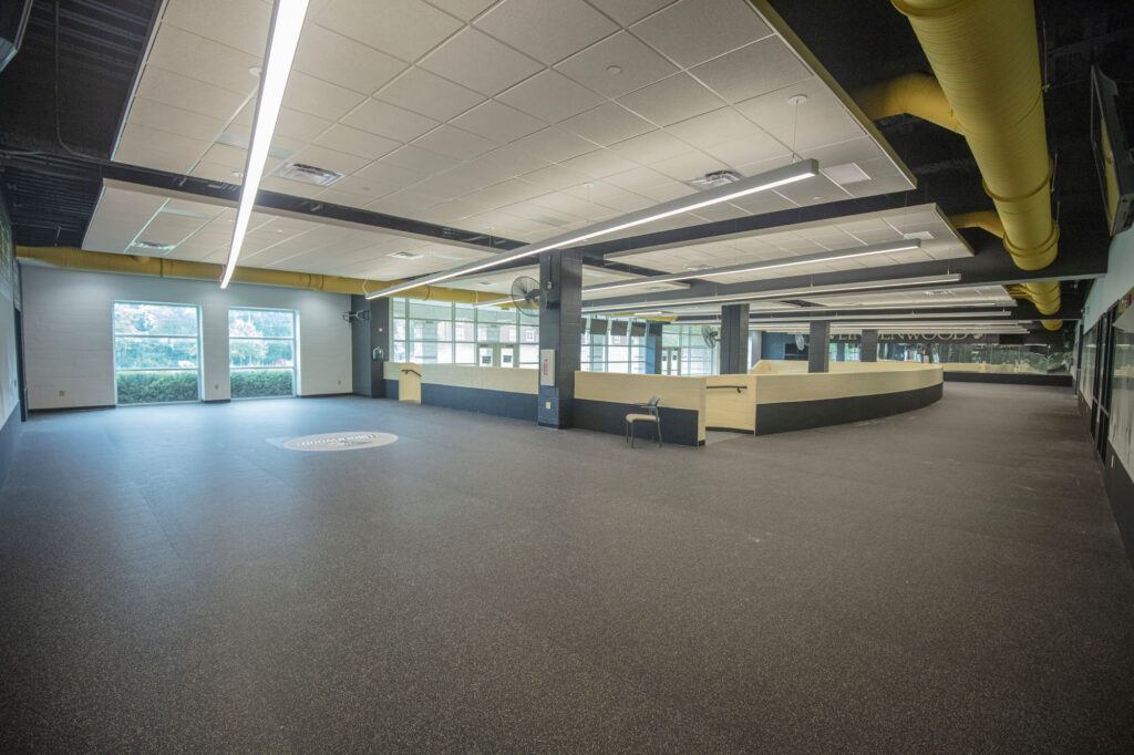 A spacious, empty room with large windows, yellow ceiling pipes, and a central curved wooden desk area. The floor is dark and textured, and fluorescent lights line the ceiling. A single chair sits near the desk.