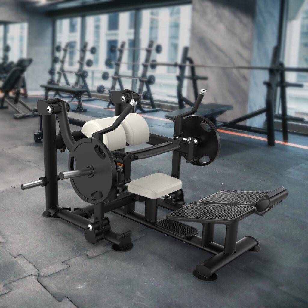 A black and white gym machine with padded seats and footrests stands on a rubber floor in a spacious gym with large windows and various weights and equipment in the background.