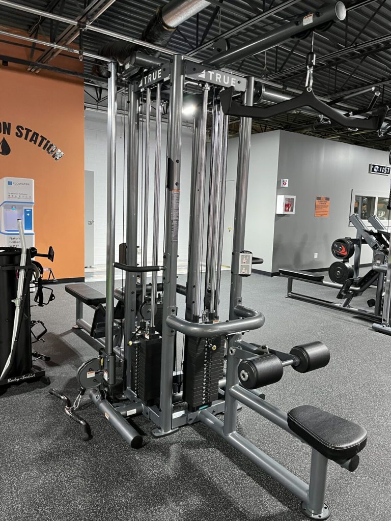 A multi-station gym machine with cables, adjustable weights, and various attachments sits in a spacious gym with gray flooring and walls. Other gym equipment and an orange wall are visible in the background.