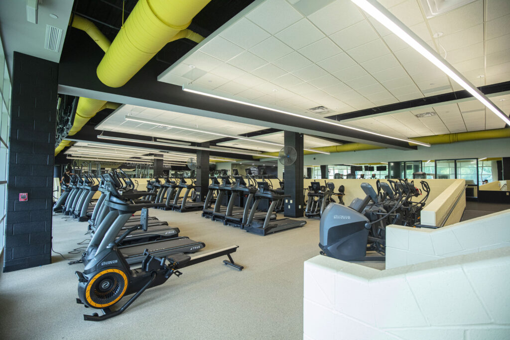 A spacious, modern gym with rows of treadmills, elliptical machines, and stationary bikes. The room has large windows, mirrors on the back wall, bright lighting, and bold yellow ventilation ducts on the ceiling.
