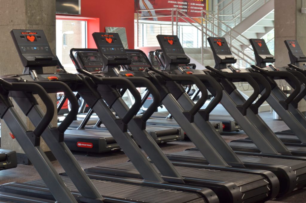 Rows of black treadmills are lined up in a gym with red and gray walls. Stairs and windows are visible in the background, and each treadmill has a digital screen. The gym is empty.