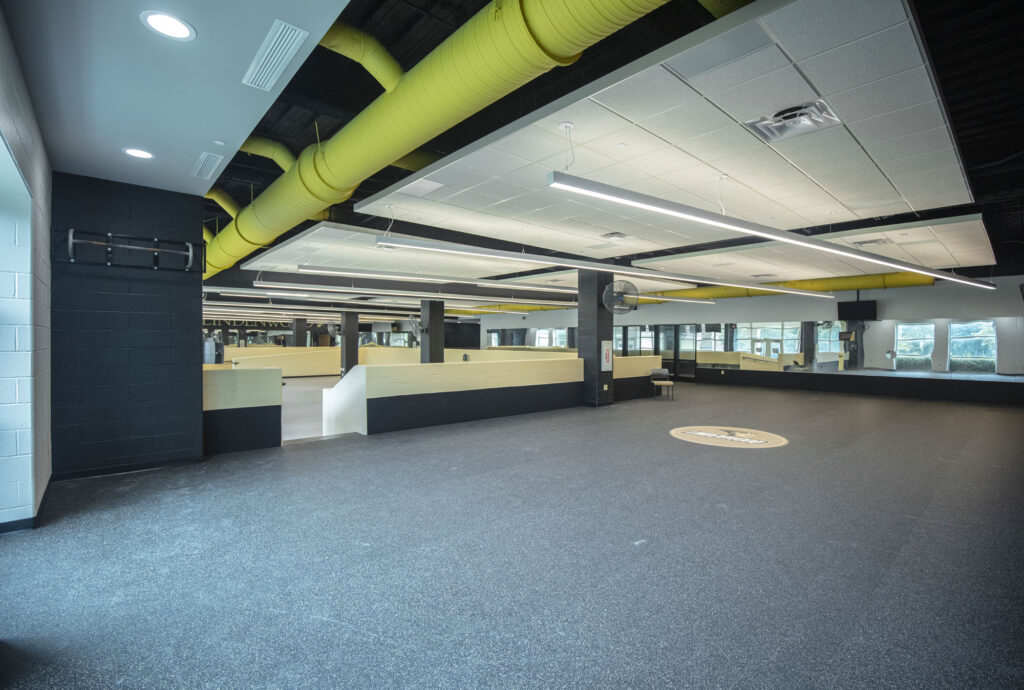 A large, open indoor area with rubber flooring, yellow overhead pipes, glass-walled sections, benches, and ample lighting, resembling a modern sports or training facility.