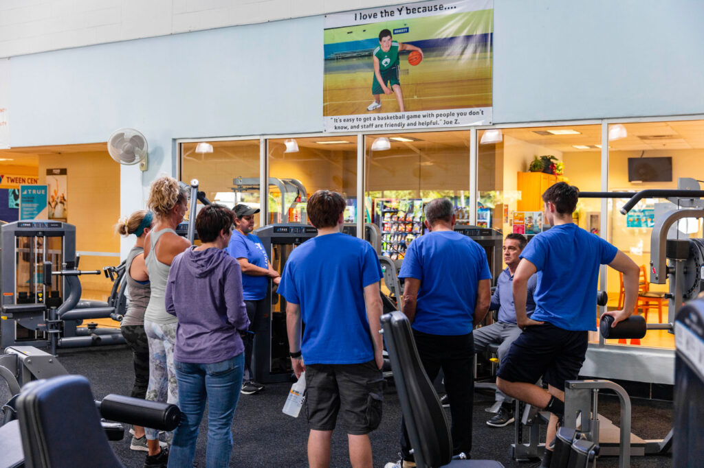 A group of people, some in blue shirts, gather around gym equipment inside a fitness center. In the background, a large poster is displayed above a windowed wall. The scene is well-lit and active.
