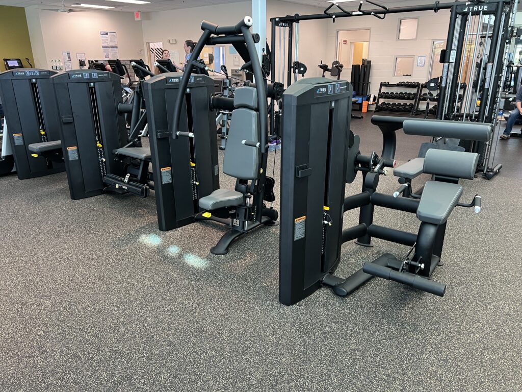 A gym with various weight machines, including leg curl, chest press, and cable machines, arranged on a rubber floor. Dumbbells are visible in the background, and the space is well-lit with a clean, organized layout.