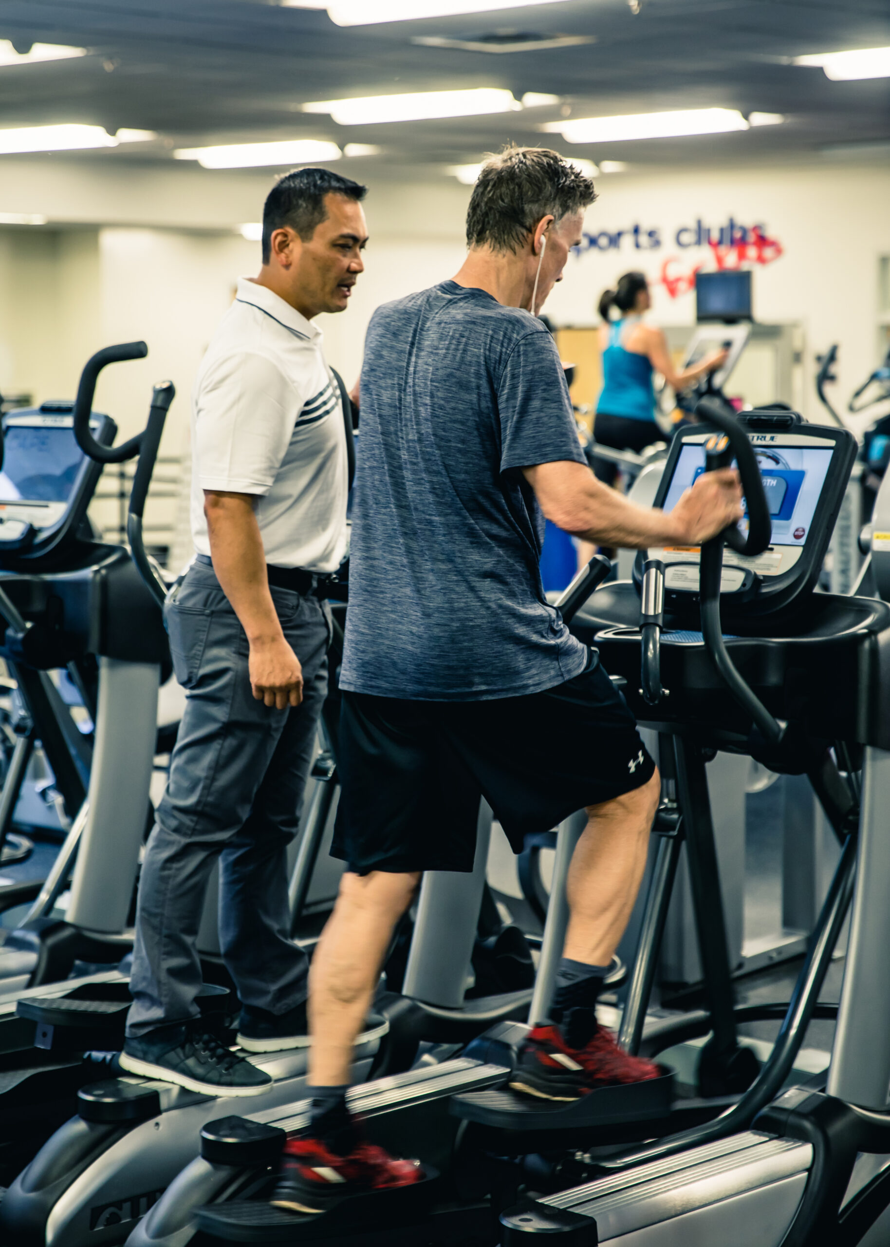 A man in athletic clothes uses an elliptical machine at a gym while another man, wearing business-casual attire, stands beside him, appearing to offer guidance. Other people exercise in the background.