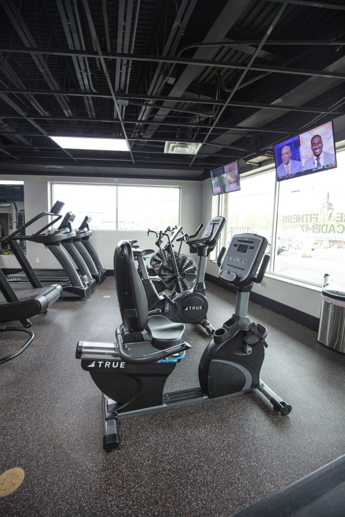 A modern gym with treadmills, stationary bikes, and cardio machines on a speckled floor. Large windows provide natural light, and two TVs are mounted on the wall showing news anchors.