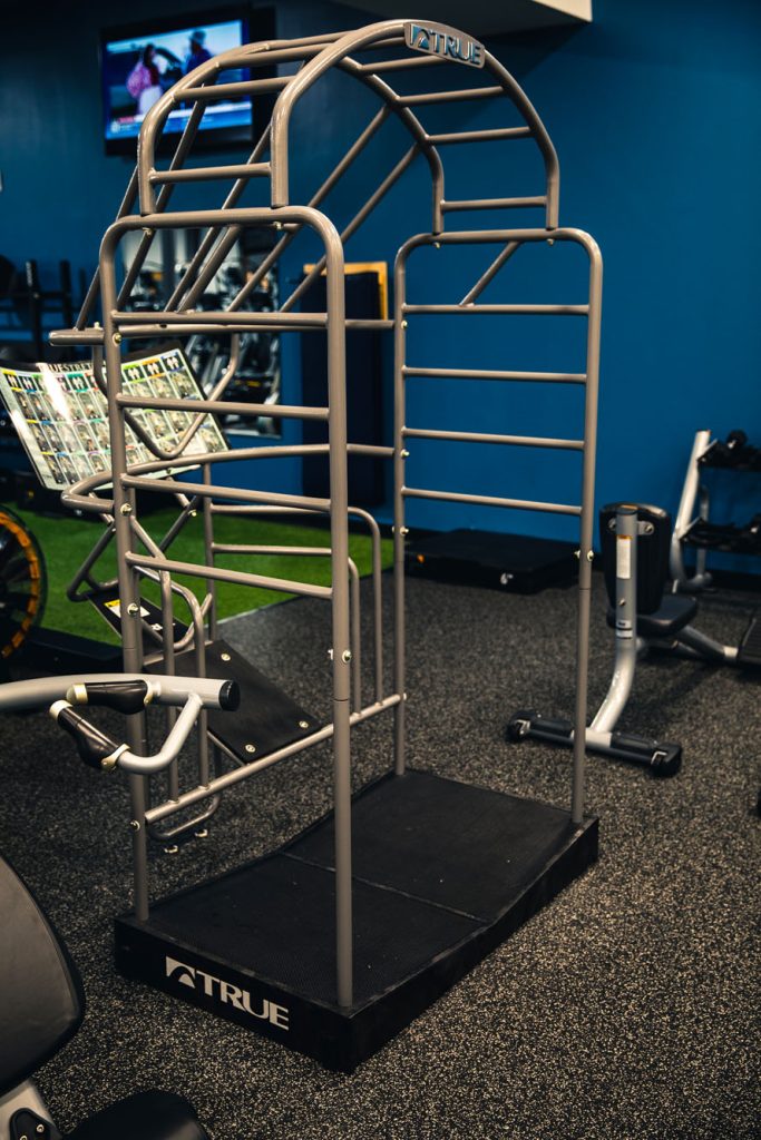 A metal exercise station with parallel bars and an arch, set on a black platform in a gym with blue walls and fitness equipment in the background.