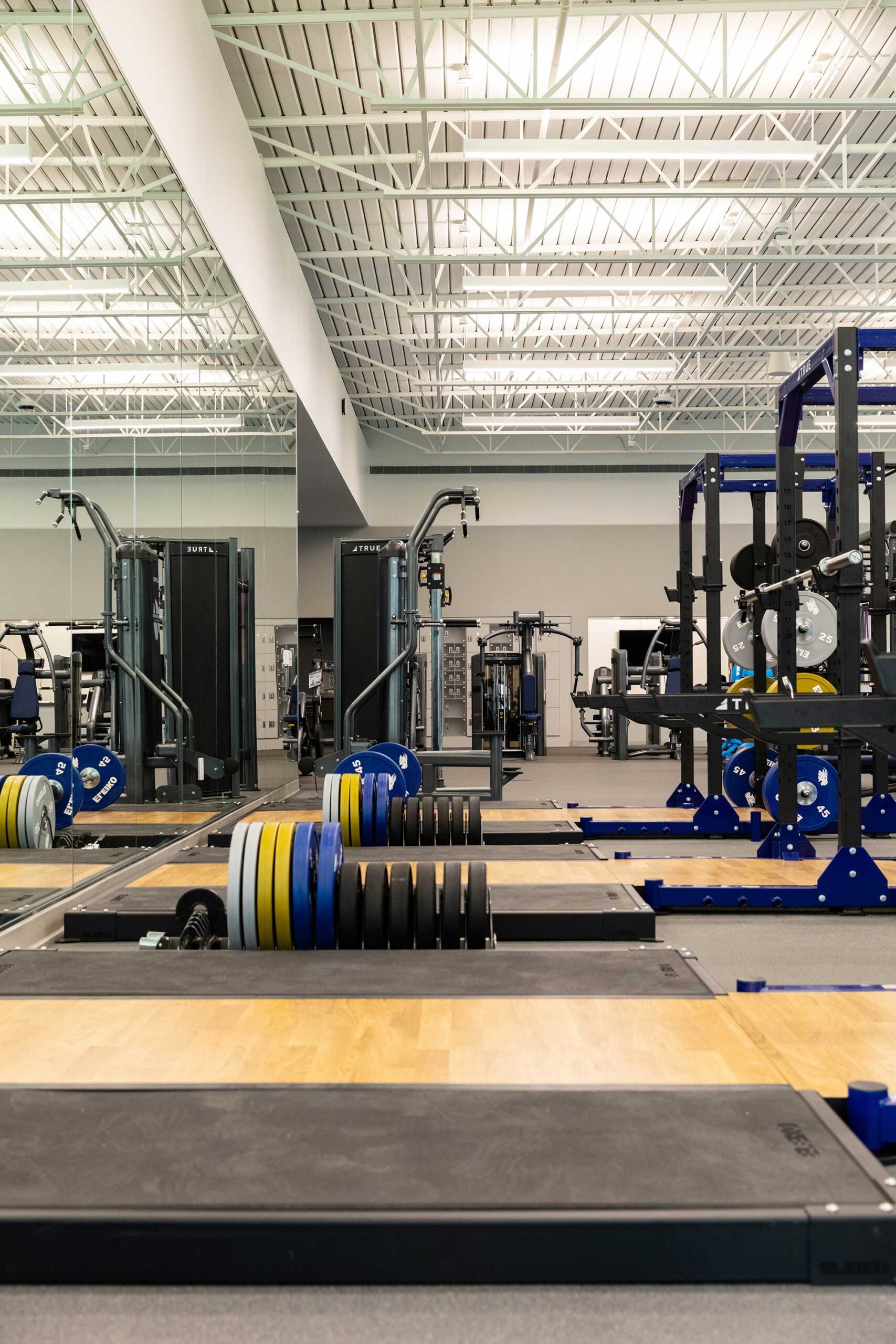 A modern gym with weightlifting platforms, barbells loaded with colorful weight plates, squat racks, and various strength-training machines under bright overhead lights.