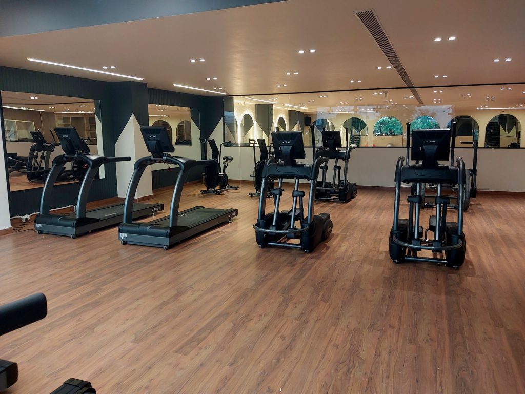 A spacious gym with wooden flooring, featuring treadmills, elliptical machines, and stationary bikes, positioned in front of large mirrors and windows that let in natural light.