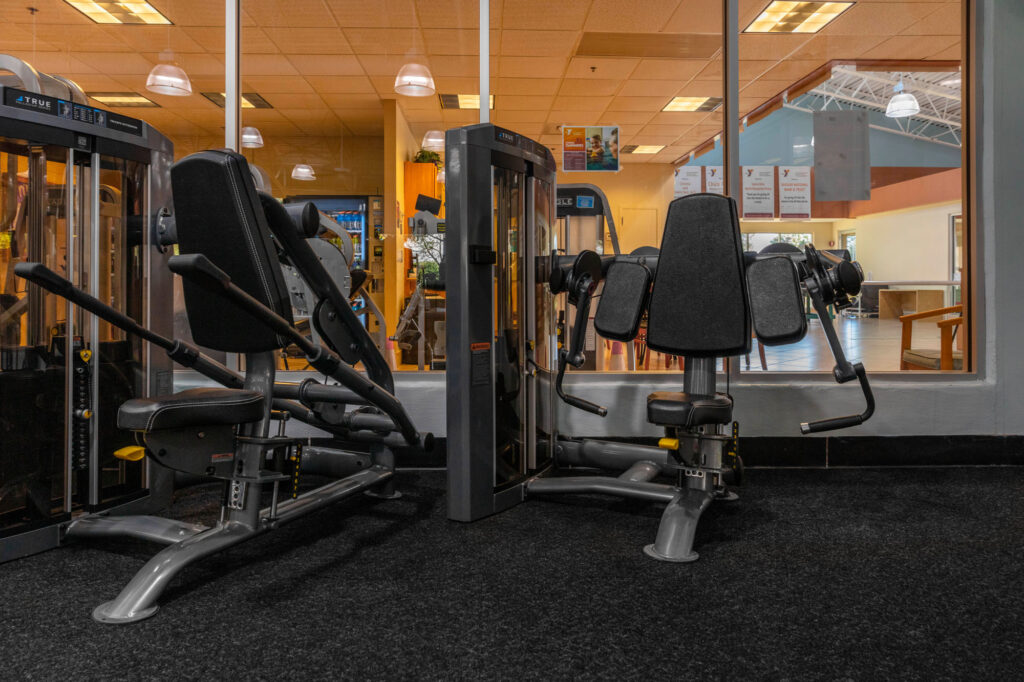 Two black gym machines with padded seats and backrests are positioned side by side on a black carpet in front of large windows, revealing a brightly lit interior with more gym equipment and wooden furniture in the background.