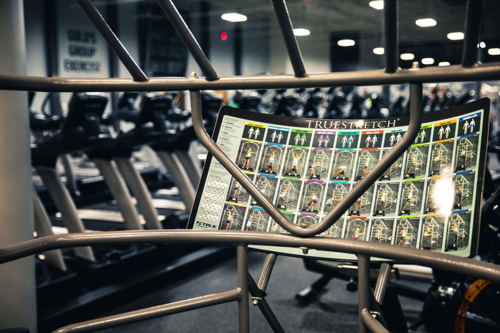 A stretching guide labeled TRUESTRETCH is attached to metal bars in a gym, with treadmills and other exercise equipment visible in the blurred background.