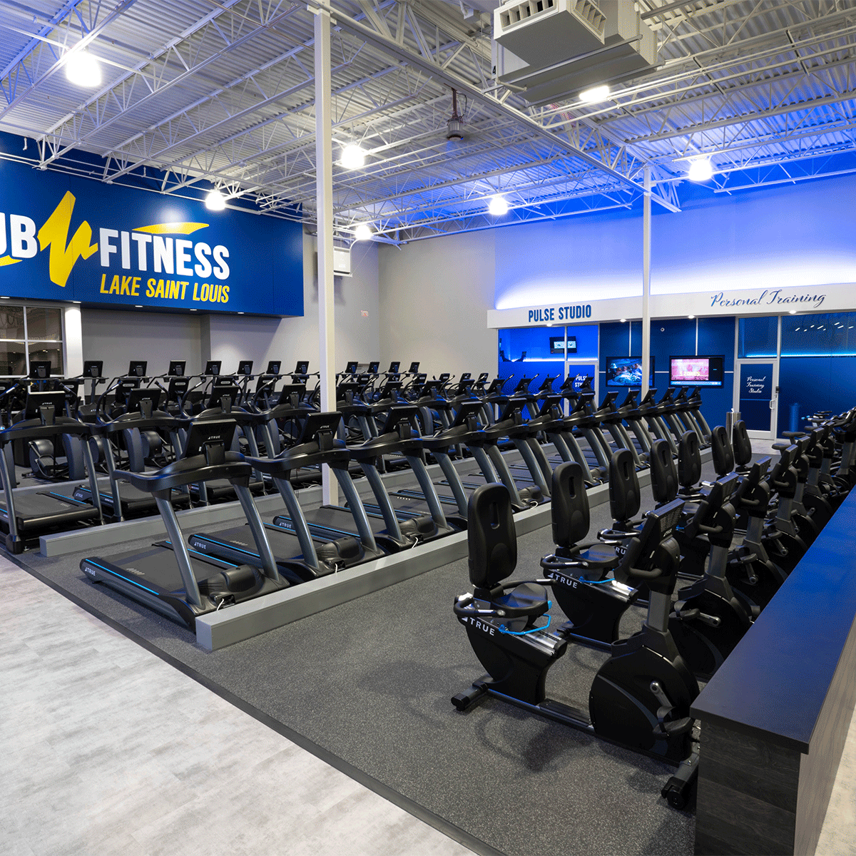 A spacious, brightly lit gym features rows of treadmills, stationary bikes, and exercise equipment. Signs read Club Fitness Lake Saint Louis, Pulse Studio, and Personal Training. The gym appears clean and modern.