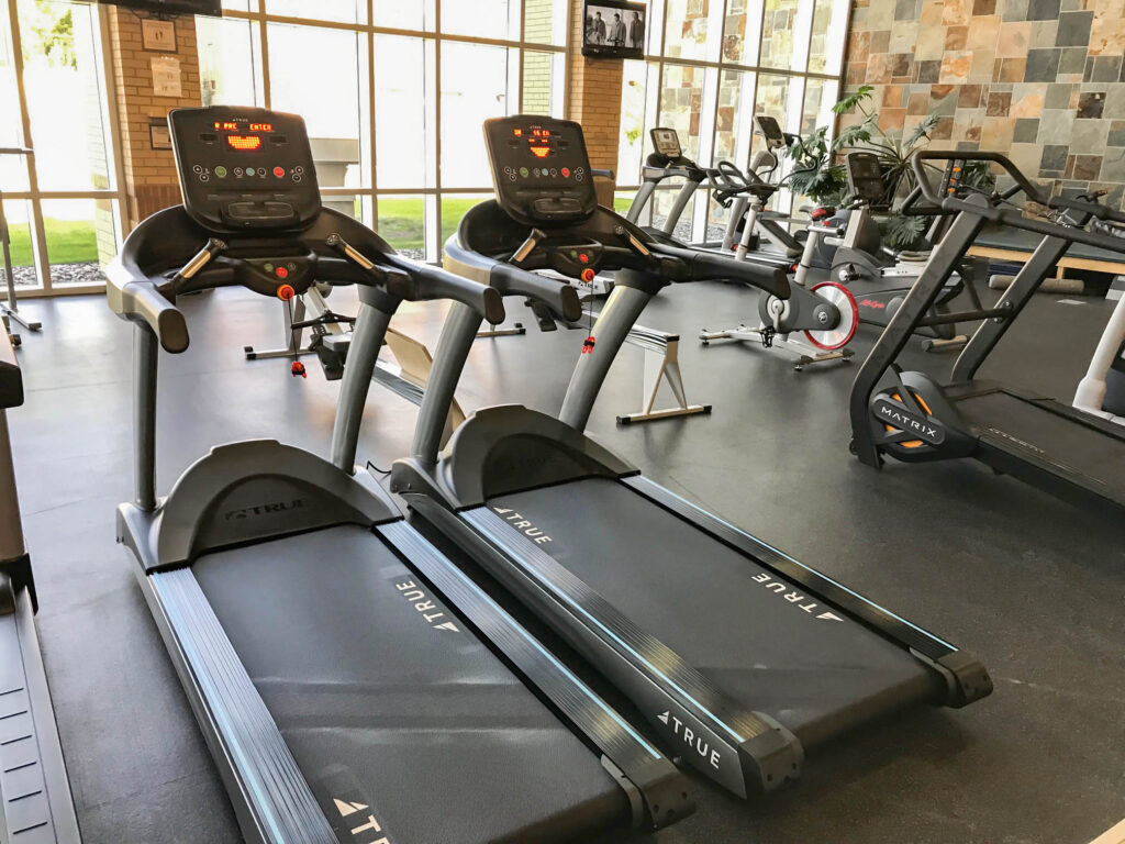 Two treadmills in a modern gym with large windows, natural light, and various exercise equipment including stationary bikes and weight machines in the background.
