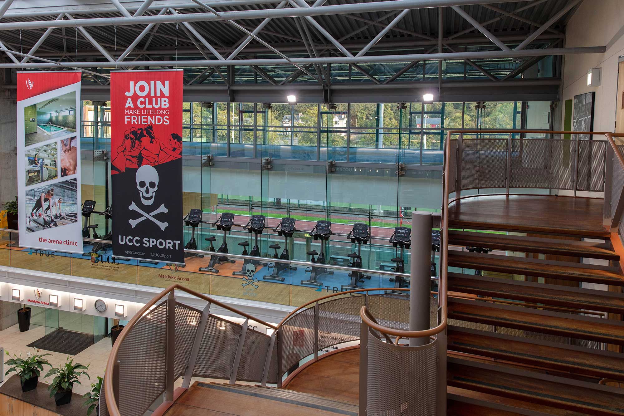 Indoor view of a modern gym with rowing machines facing large windows. Two banners hang in front, one red with text Join a club, make lifelong friends, and another black and red for UCC Sport with a skull and crossed oars.
