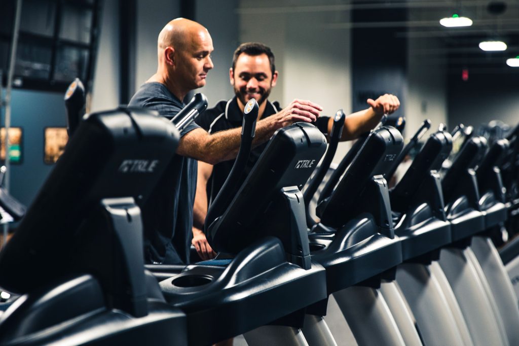 Two men stand side by side among a row of treadmills in a gym. One man is adjusting the controls on a treadmill while the other smiles, appearing to give guidance or instruction.