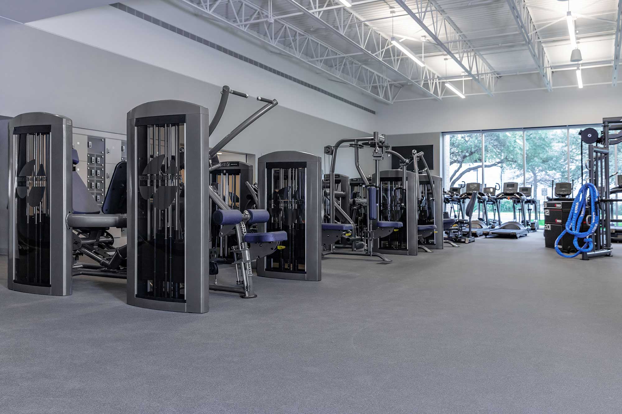 A spacious, modern gym with various strength training machines, exercise bikes, and treadmills. The room has high ceilings, large windows letting in natural light, and a clean, uncluttered appearance.