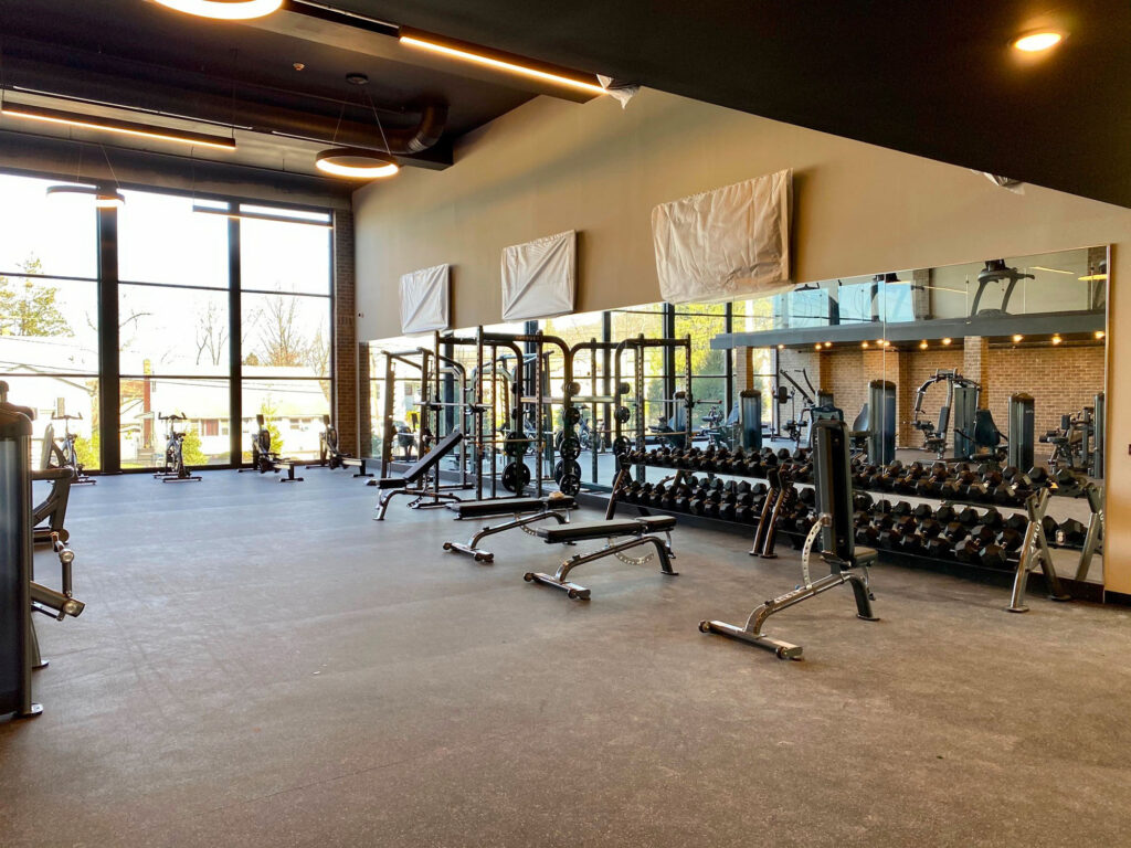 Spacious gym with large windows, weight racks, benches, and various exercise machines. The room has mirrored walls and natural light. No people present.
