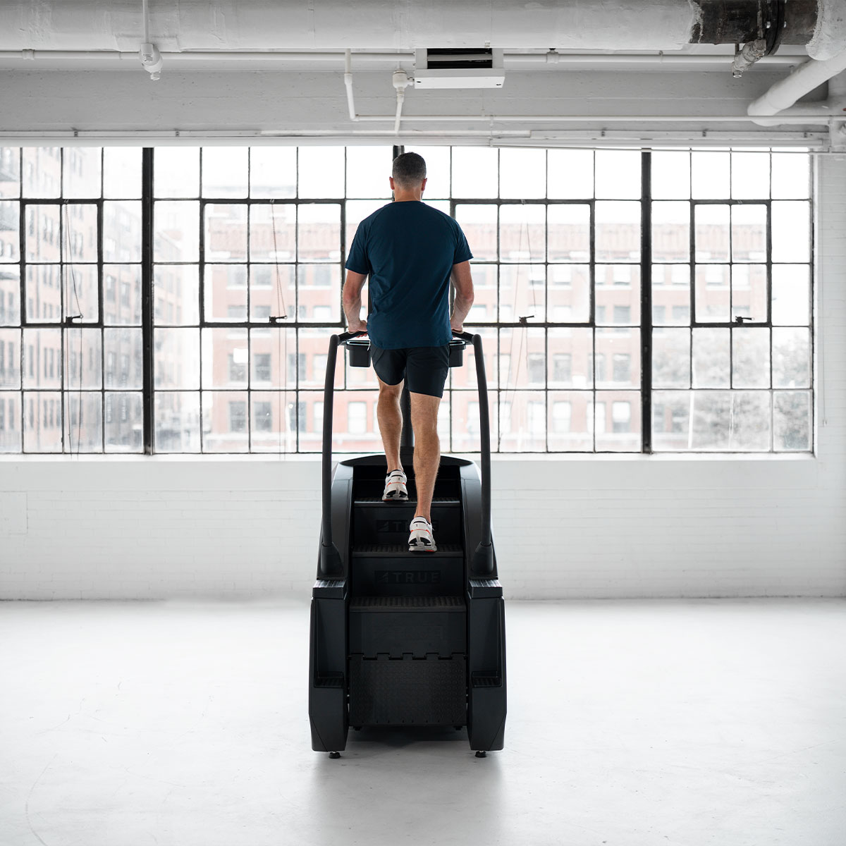 A person in athletic wear uses a stair-climbing machine in a bright, spacious gym with large windows overlooking city buildings.