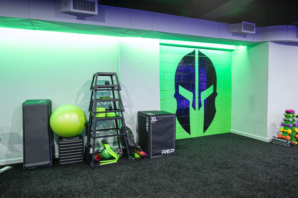 A gym corner features a green stability ball, stacked step platforms, kettlebells, a plyo box, colorful dumbbells, and a neon-lit wall with a black Spartan helmet design.