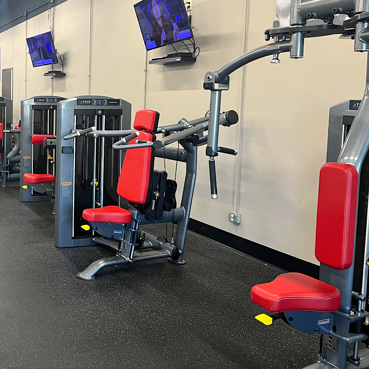 A gym with red and gray weight machines lined up against a wall. Several flat-screen TVs are mounted above the machines, and the area has black flooring and beige walls. The gym appears clean and empty.