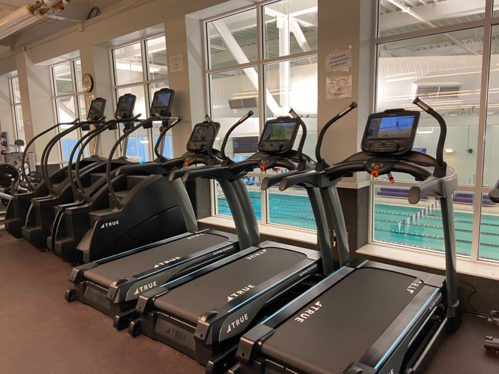 A row of treadmills and elliptical machines faces large windows overlooking an indoor swimming pool with clear blue lanes inside a well-lit fitness center.