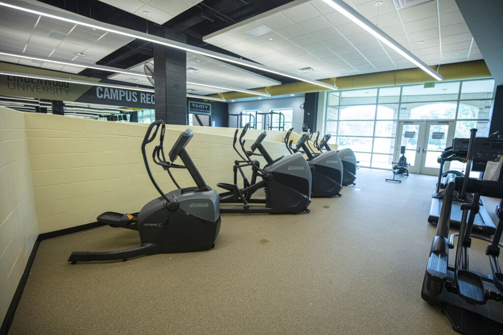 Three elliptical machines are lined up against a low beige wall in a bright gym with large windows and mirrors. The gym also features cardio equipment and has light-colored flooring and ceiling panels.