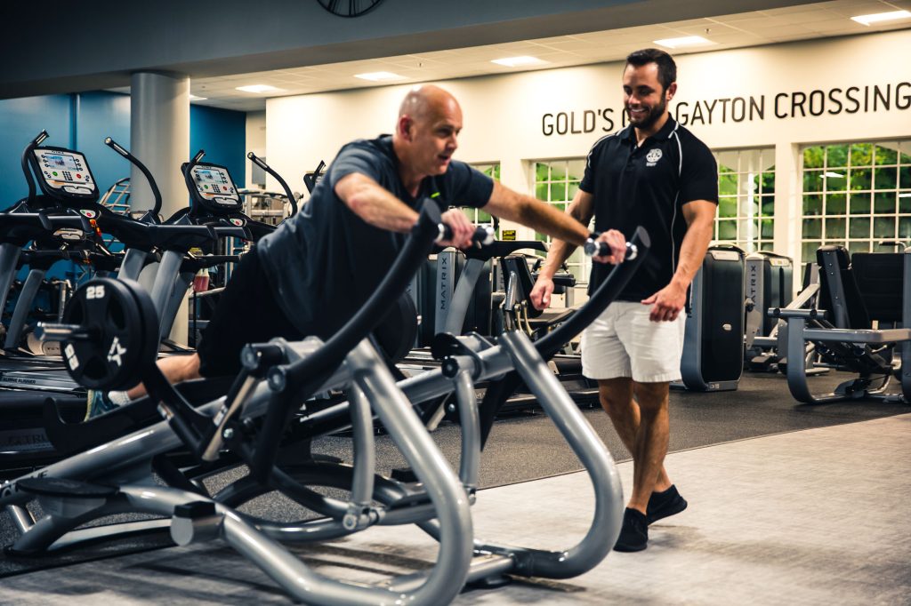 A man works out on a rowing machine in a gym while a trainer in a black shirt and shorts stands nearby, smiling and offering encouragement. Treadmills and gym equipment are visible in the background.