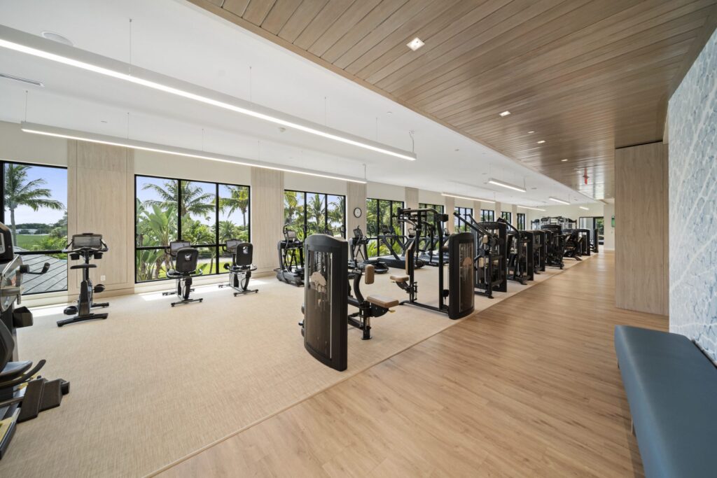 Spacious, modern gym with wood floors and ceiling, large windows with views of palm trees, rows of cardio and strength machines, and natural light filling the room.