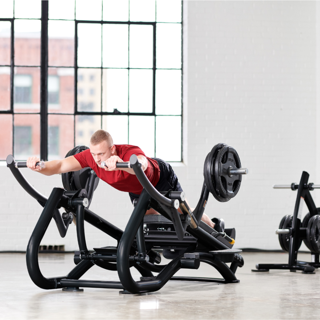 A man in a red shirt uses a weight machine, lying face down and pushing handles forward in a bright gym with large windows and gym equipment in the background.