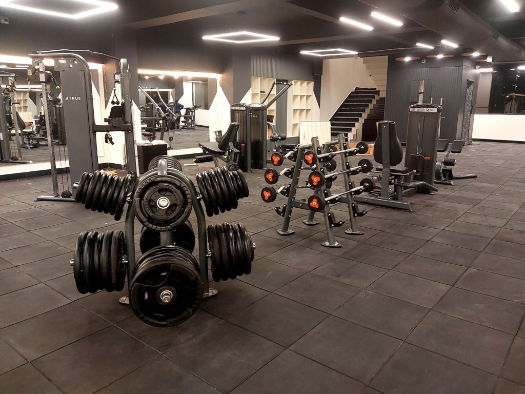 Modern gym interior with weight racks, dumbbells, various exercise machines, and rubber flooring. The space is well-lit with ceiling lights and features mirrors and stairs in the background. The gym is empty.