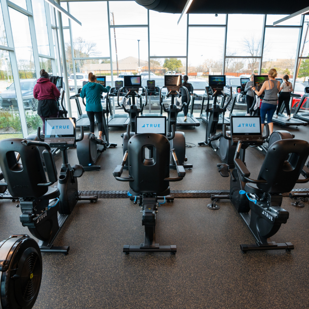 People work out on cardio machines, such as exercise bikes and treadmills, in a bright, modern gym with large windows and outside views. Several empty stationary bikes are in the foreground.