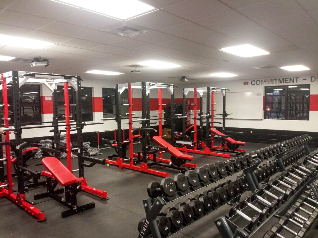 A gym with rows of dumbbells, red and black weight benches, and squat racks. The room has bright lighting, mirrors on the walls, and motivational words printed near the ceiling.