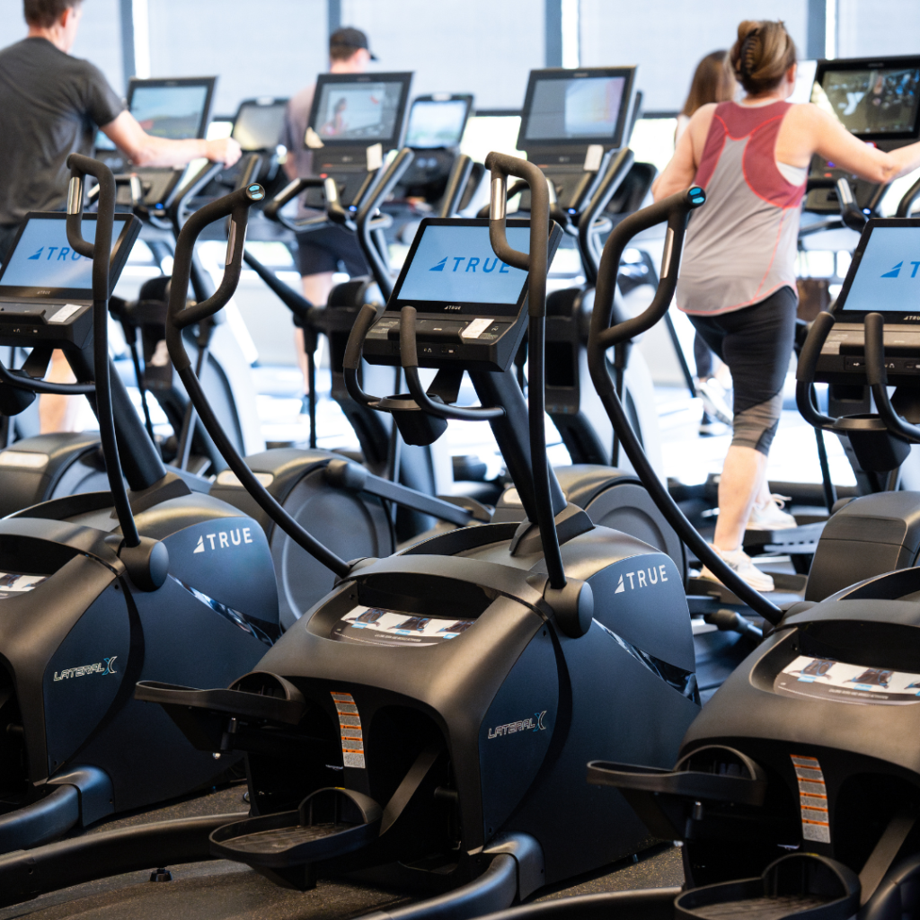 People exercising on elliptical machines in a bright gym, with several screens and rows of equipment visible. Most people are wearing athletic clothing and facing away from the camera.