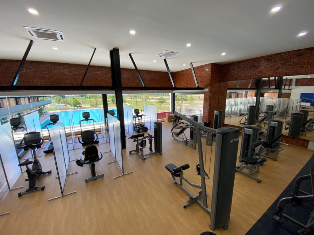 A modern gym with various exercise machines arranged on a wooden floor, large windows overlooking an outdoor pool, and clear dividers between equipment. The space is well-lit with ceiling lights.
