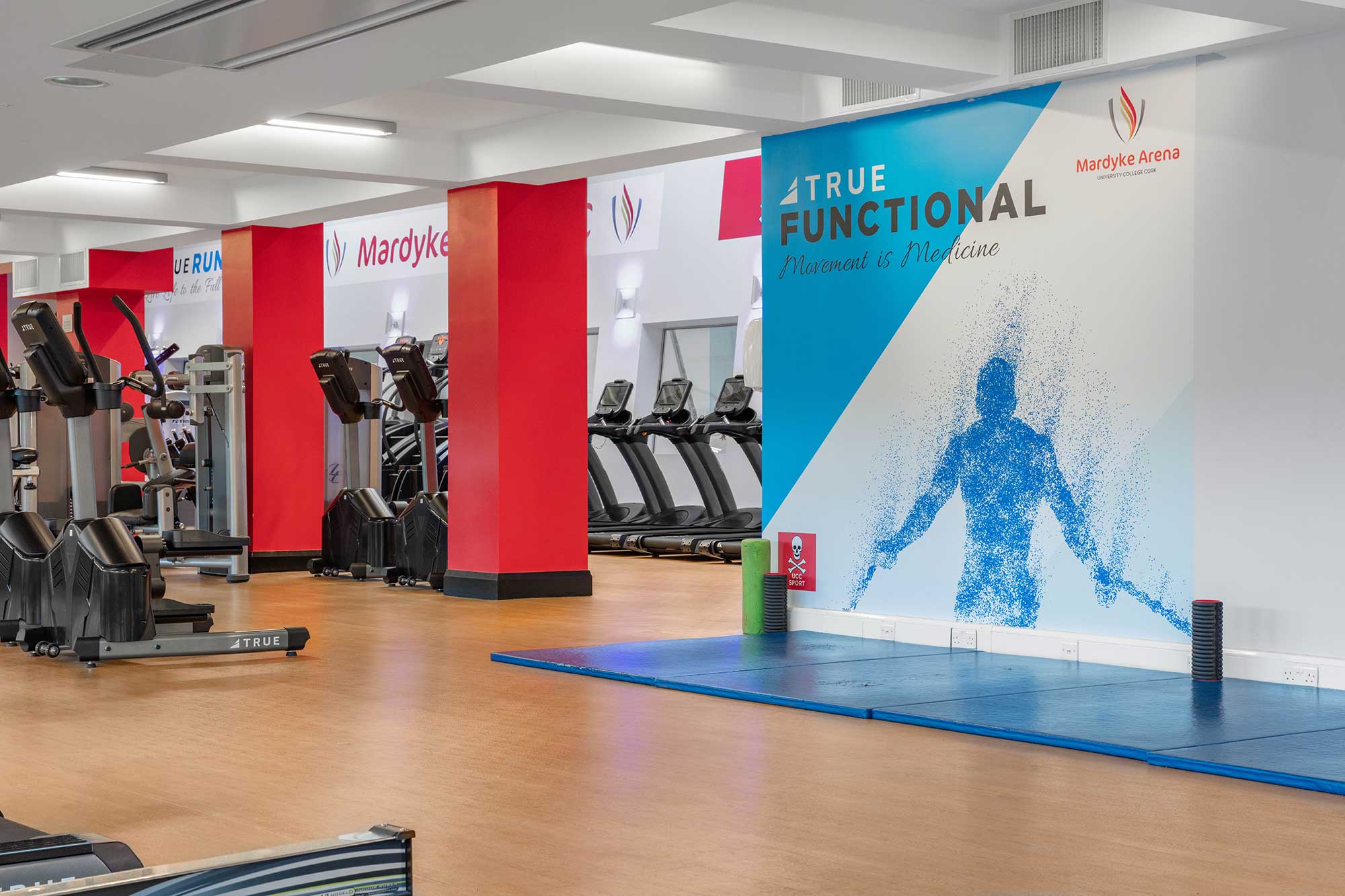 A modern gym featuring treadmills, exercise bikes, and a blue mat area with a “True Functional Movement & Medicine” sign on the wall. The space is well-lit with red and white accents.
