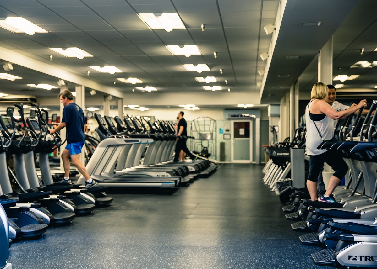 People using exercise machines in a spacious, well-lit gym with rows of treadmills and elliptical trainers; the room has a clean, modern appearance.