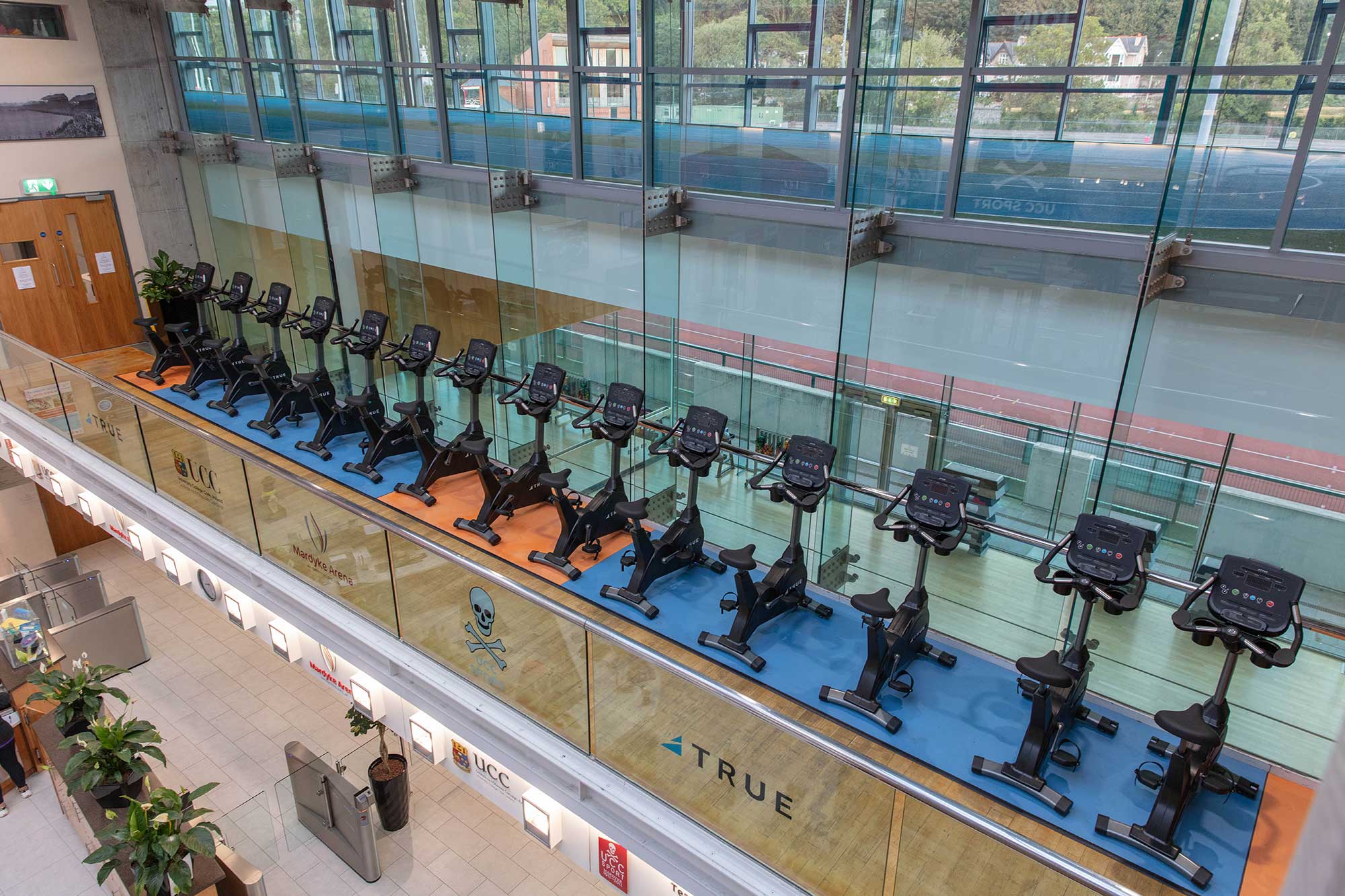 A row of stationary exercise bikes is lined up on an indoor balcony with glass walls, overlooking a running track below. The area is brightly lit with natural light from large windows.